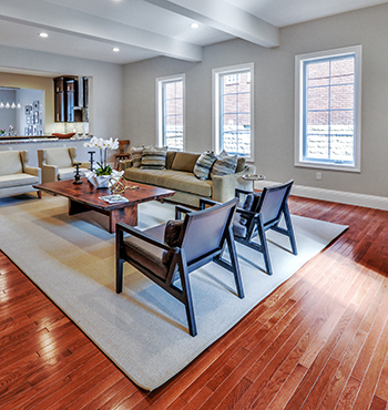 Living room with wood floors, a white rug, and wooden furniture. Open to the kitchen in the background.