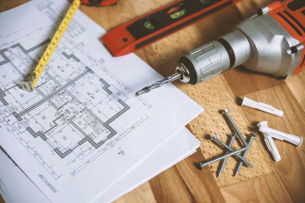 Blue prints of house construction project on table next to nails, screws, hand drills, and ruler.