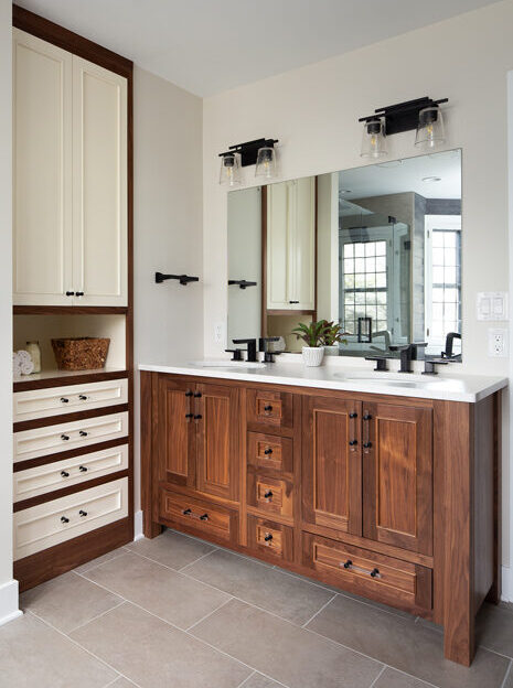 Traditional bathroom remodel with woodgrain paneled cabinets, stone dual-vanity countertop, matte black faucets, and large gray floor tiles.